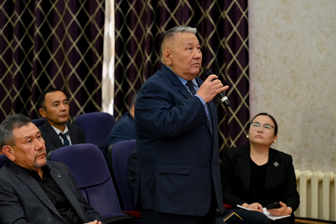 Микрофон ұстап, сөйлеп тұрған костюм киген ер адам және оны тыңдап отырған адамдар.