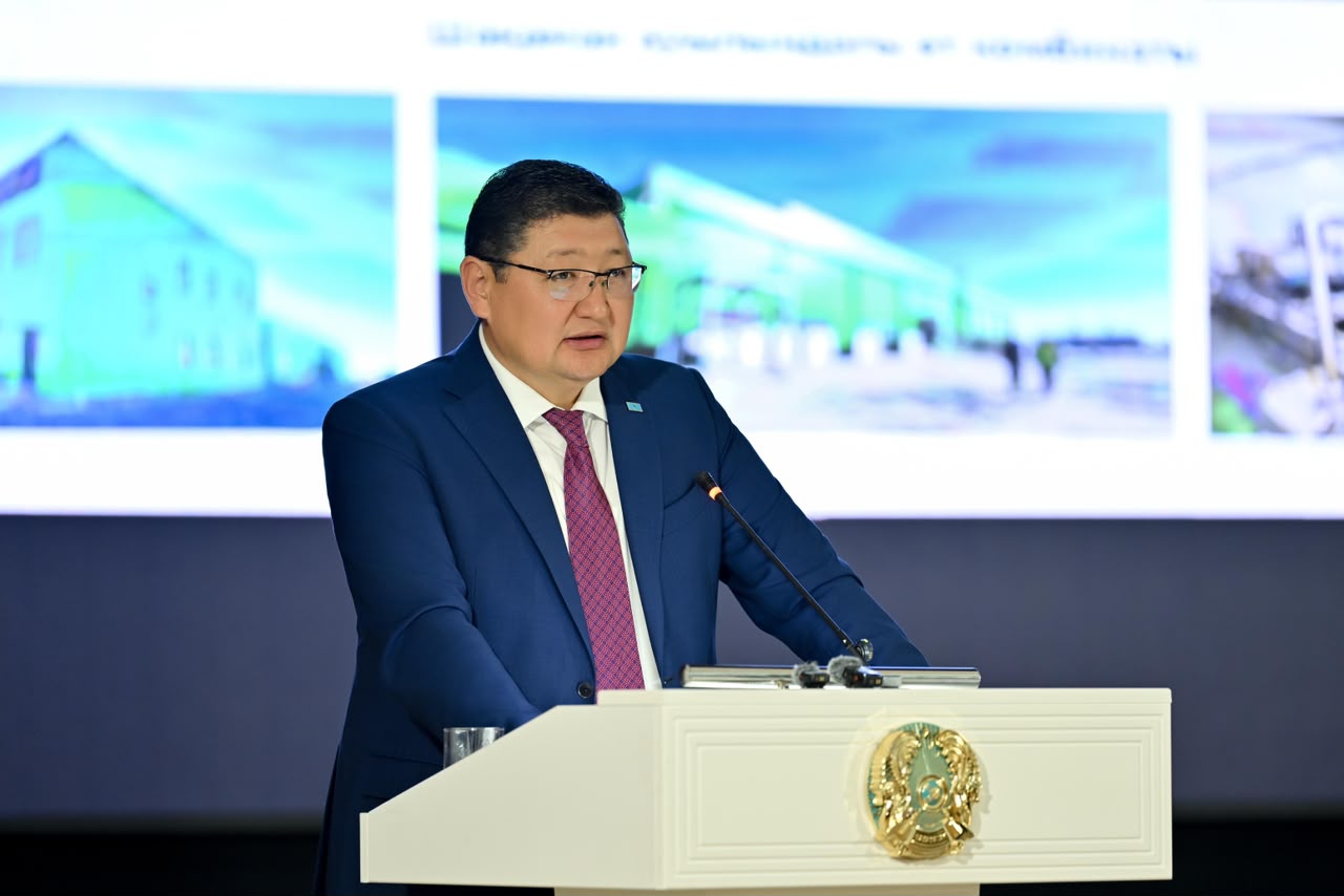 Көк костюм киген ер адам трибунада сөйлеп тұр.