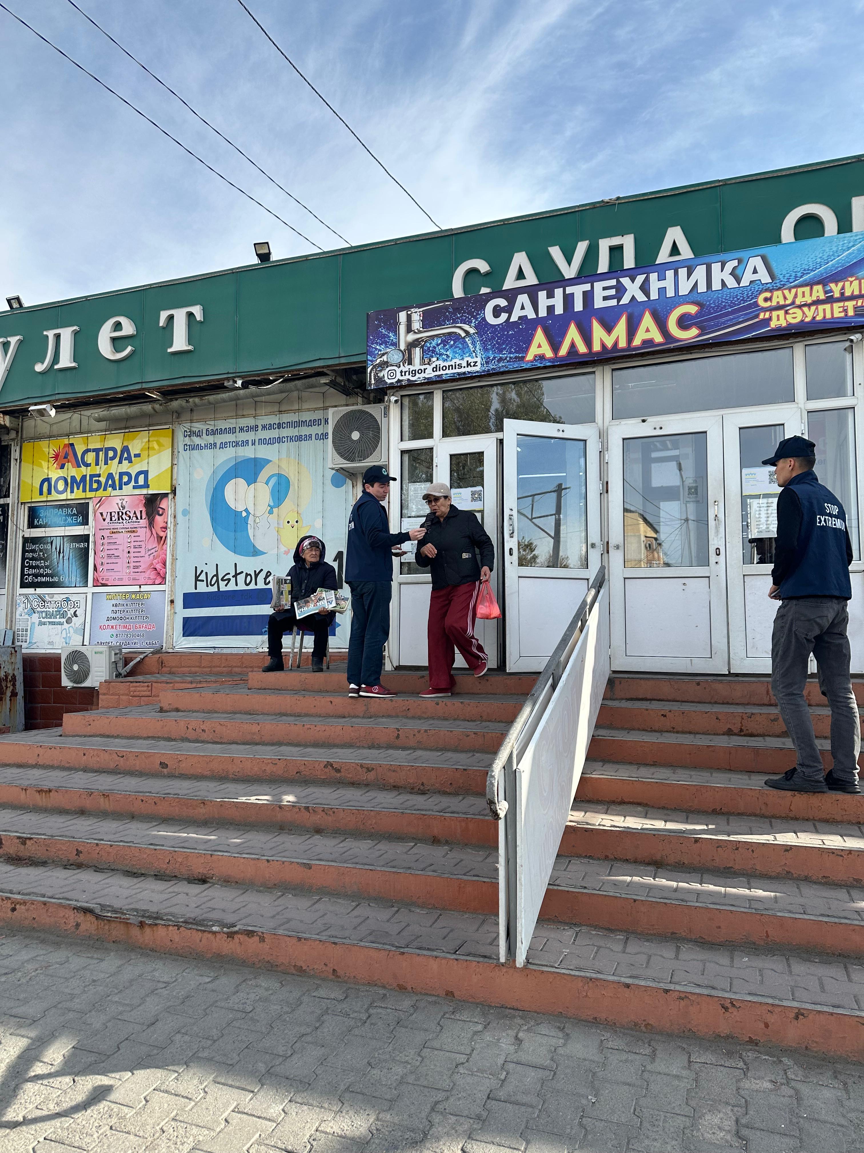 Дүкеннің кіреберісіндегі адамдар және сауда орталығының жарнамалық белгілері.