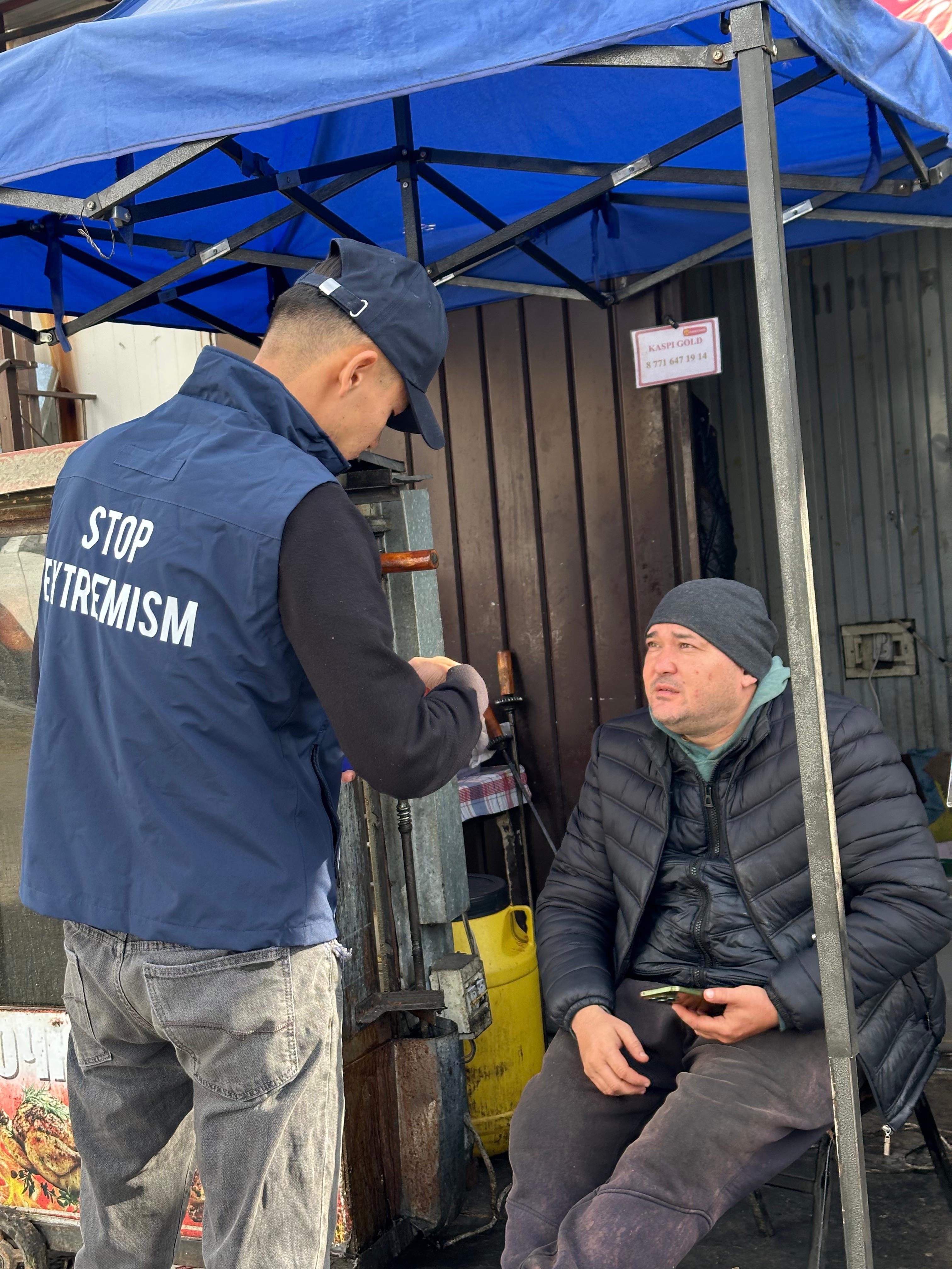 «Stop Extremism» жазуы бар жилет киген адамның ер адаммен сөйлесіп тұрғаны.
