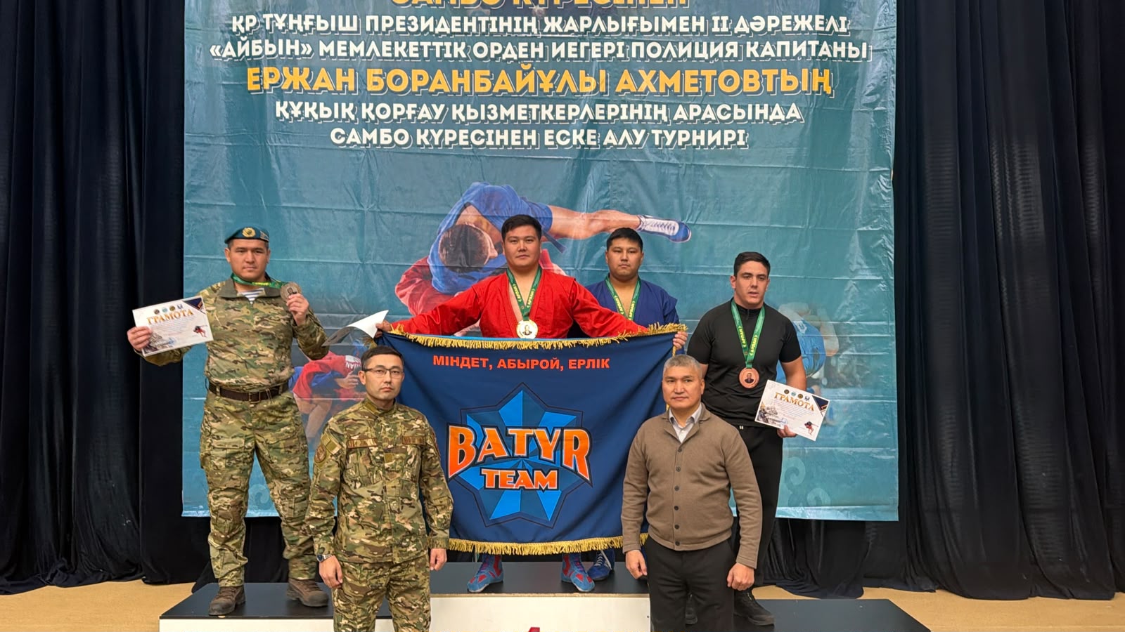 Группа мужчин с медалями и грамотами позирует с флагом «BATYR TEAM» на фоне баннера турнира по самбо.